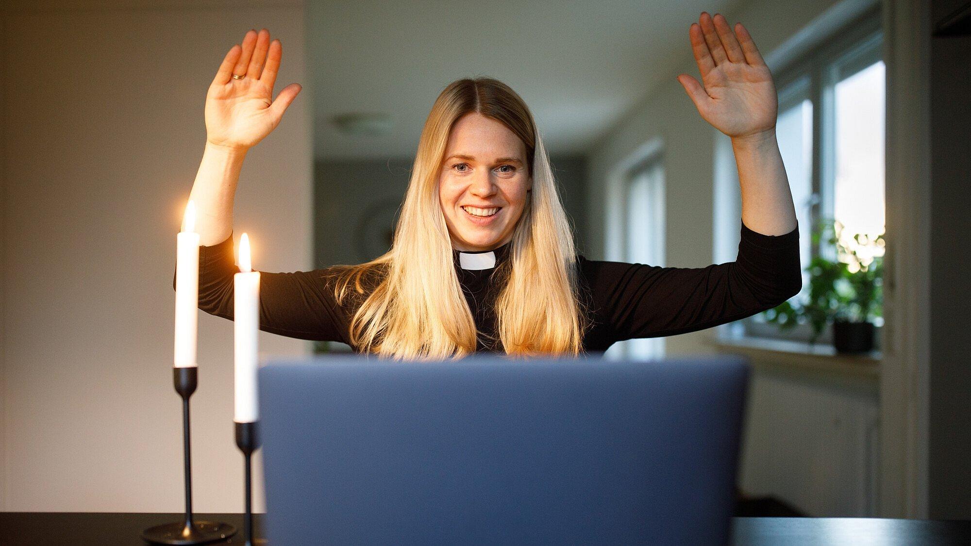 Eine Pfarrerin hält ihre Arme zum Segen hoch während eines Online-Gottesdienstes und schaut in eine Laptop.  Zwei Kerzen brenen neben ihr auf dem Tisch.E