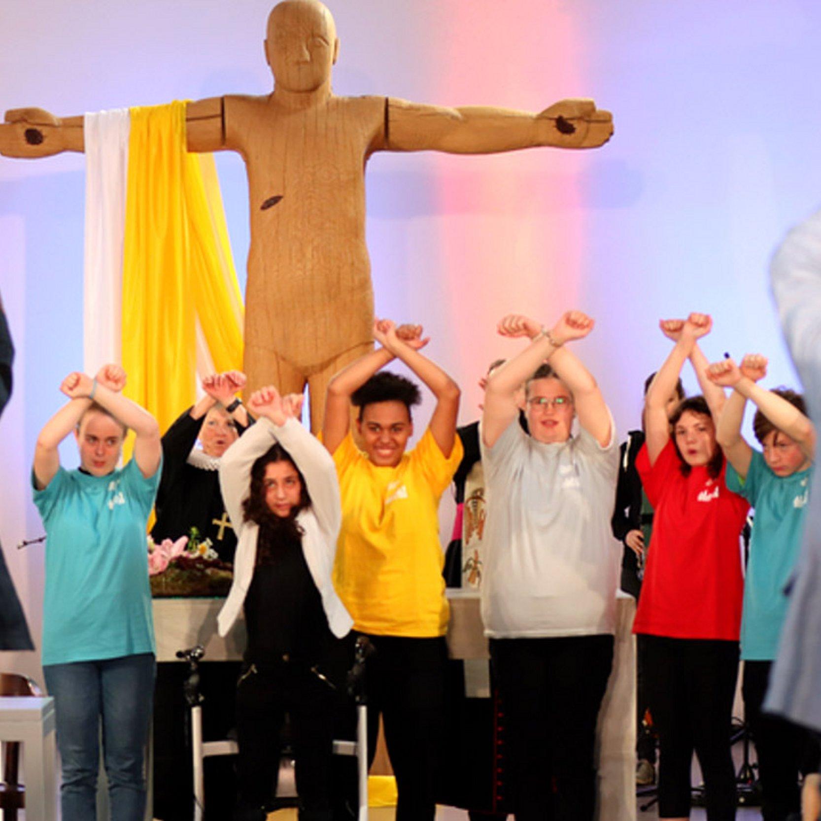 Junge Leute heben die Arme in die Höhe, dahinter eine große, hölzerne Jeusus-Statue