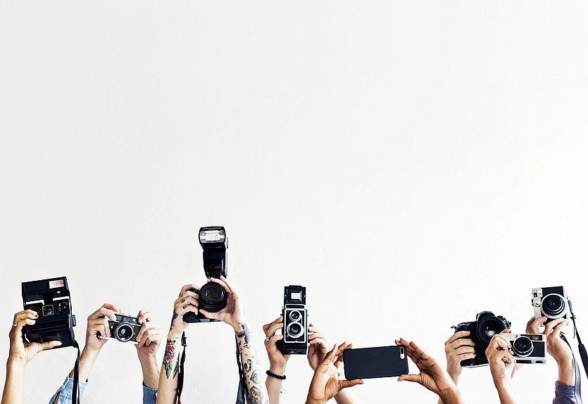 Head_News Mehrere erhobene Hände halten unterschiedliche Kameras und ein Smartphone nach oben gerichtet, als würden sie fotografieren, vor einem schlichten weißen Hintergrund.