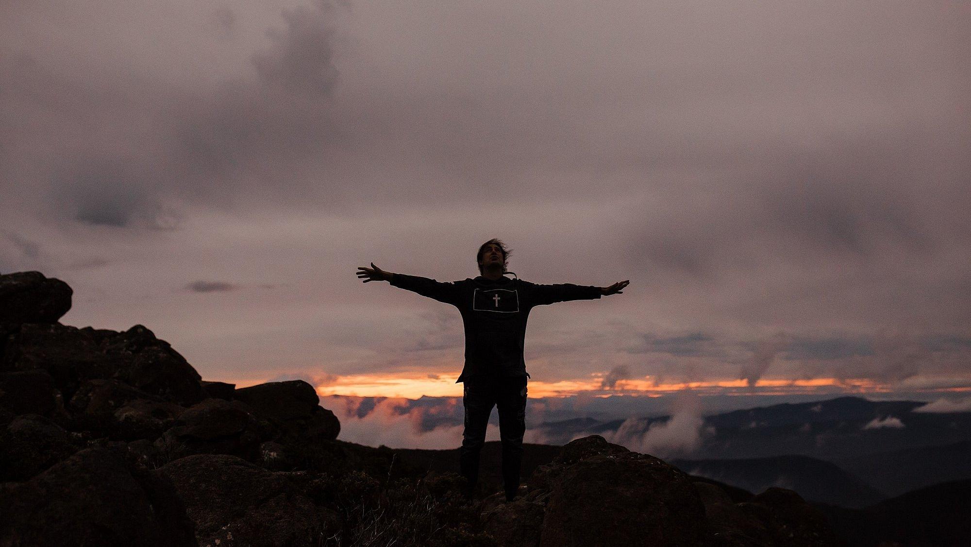 Mann steht mit ausgebreiteten Armen auf einem Berg vorm Himmel.