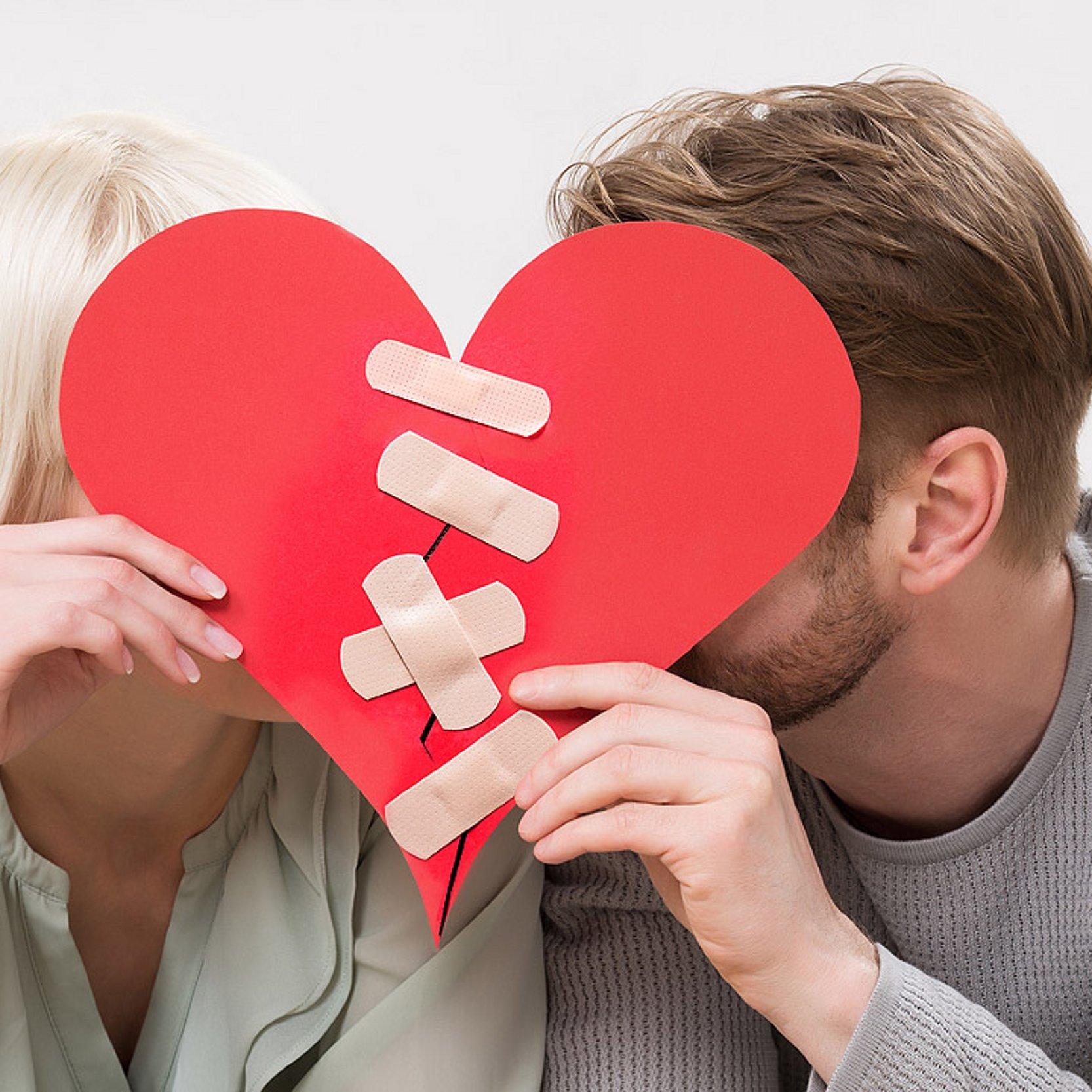 Versöhnung nach Beziehungskrise Paar hinter einem roten Pappherz, durch das ein Riss geht, der mit Wundpflastern zugeklebt wurde