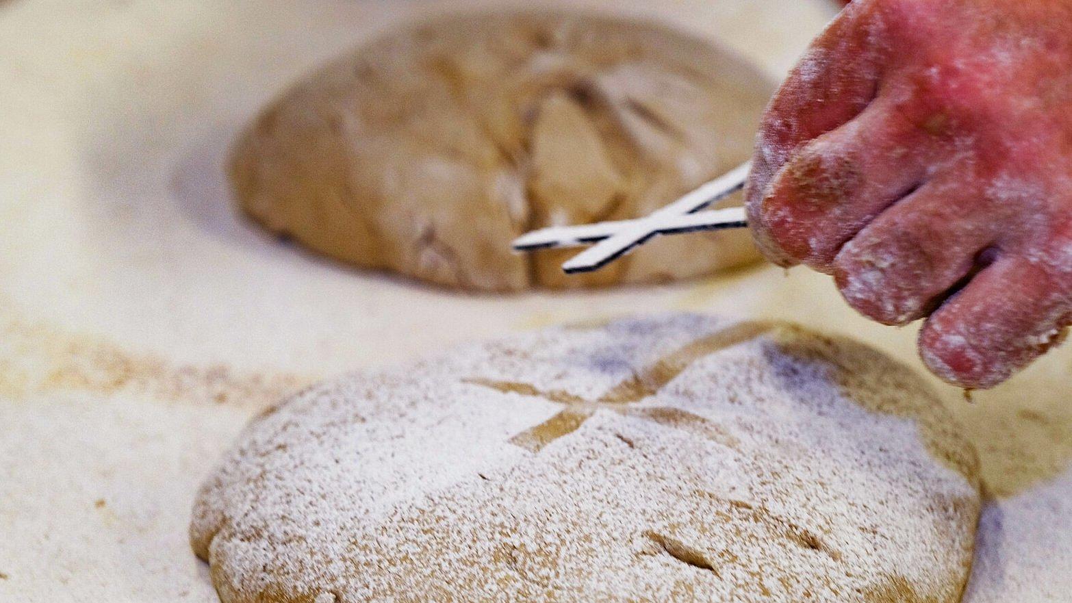 In einer Bäckerei wird Brot gebacken für die Aktion "5.000 Brote - Brot für die Welt". Im Vordergrund ein Brot und eine Hand, die ein Kreuz hält, um das Kreuz als Motiv auf das Brot zu bringen.