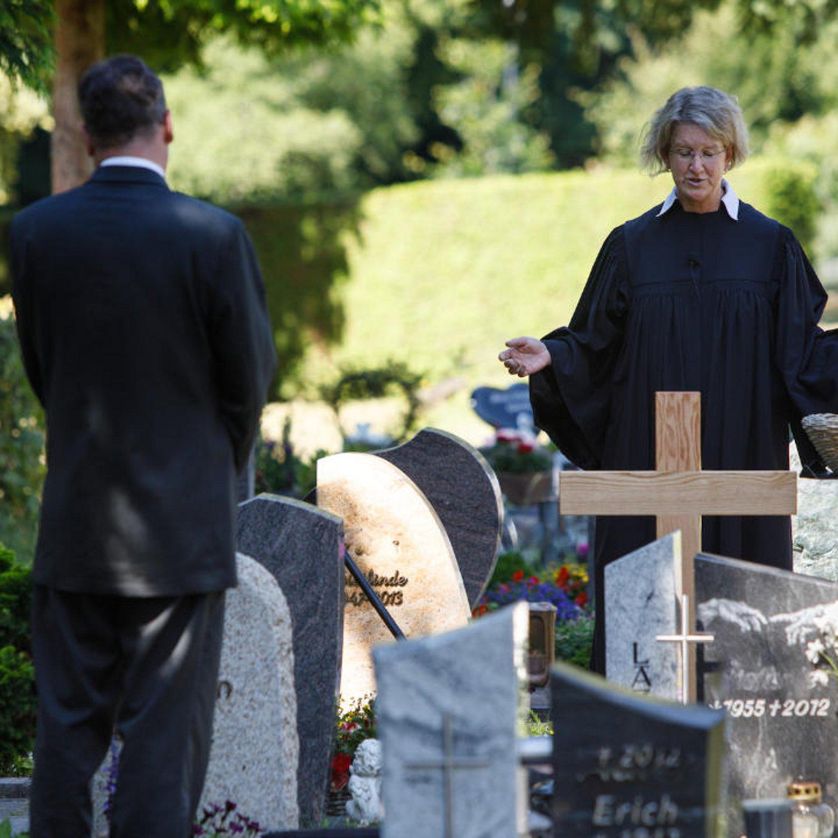 Eine evangelische Pfarrerin am Grab zwischen vielen Grabsteinen. Ein Mann steht im Vordergrund