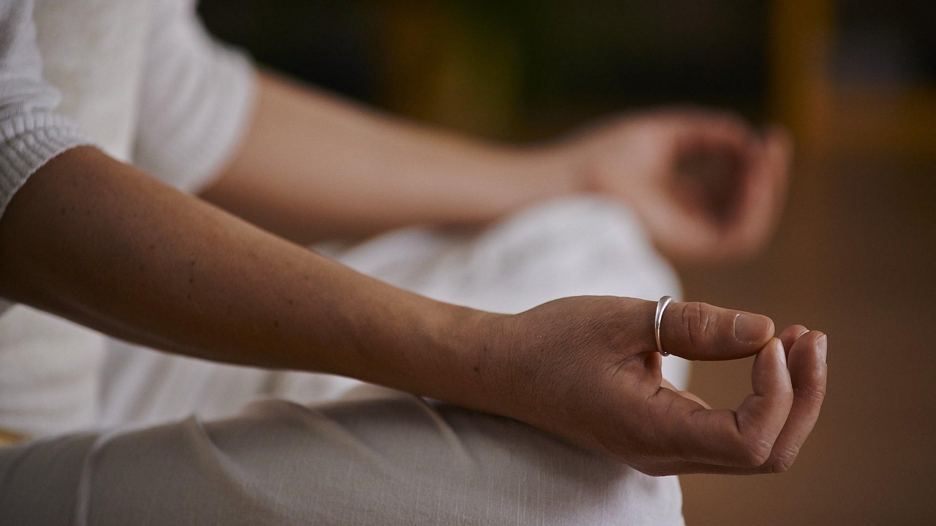 Bei einer Yoga-Übung liegen die Hände einer Person, die weiße Kleider trägt, auf ihren Knien.