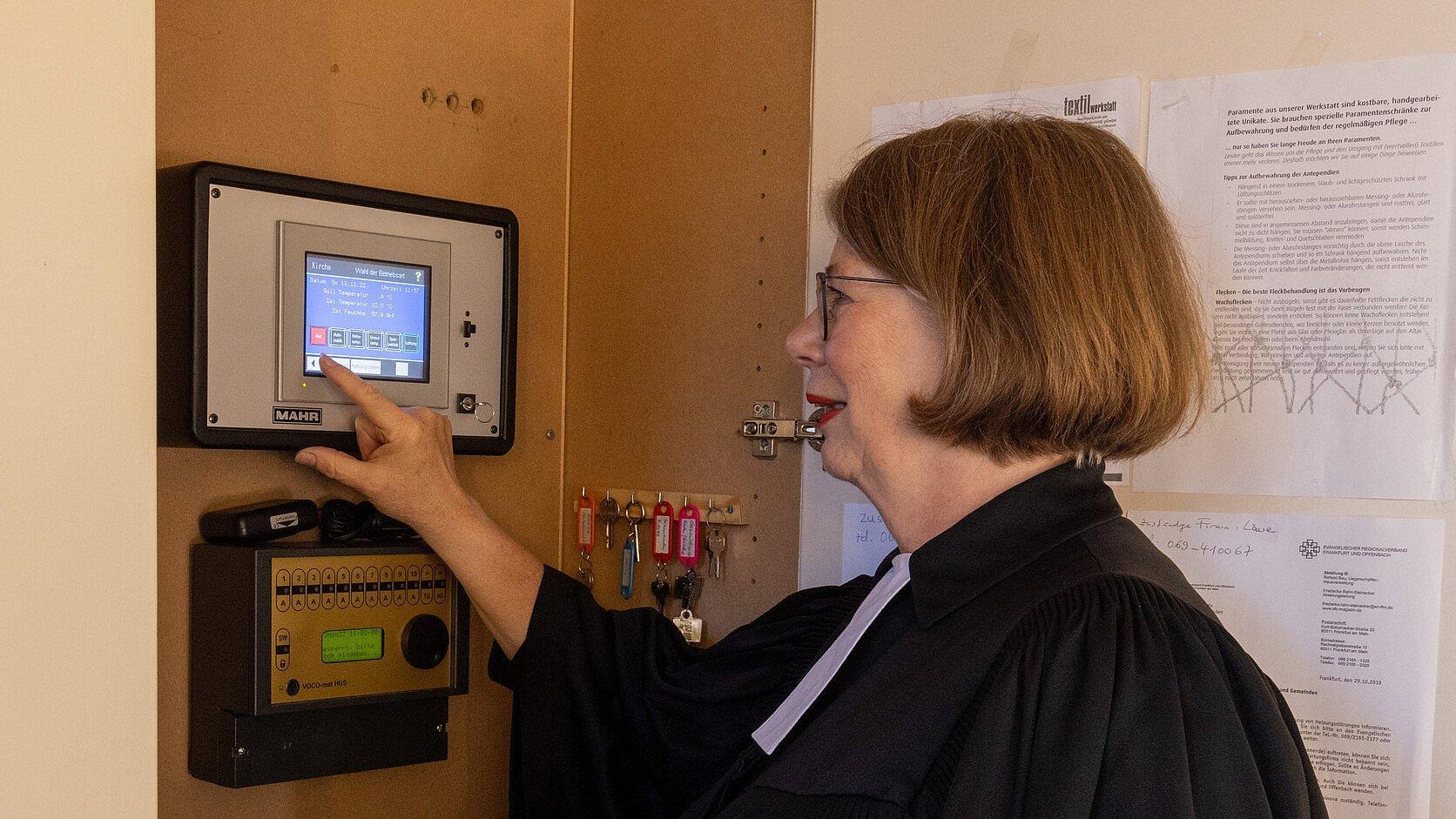 Energiesparen in der Kirche. In der Nazarethkirche in Frankfurt Eckenheim wird die Temperatur des Kircheninnenraumes über ein Thermometer gemessen und und die moderne Heizung lässt sich über ein Display steuern. Gemeindepfarrerin Christine Streck-Spahlinger zeigt die Heizungssteuerung im Kirchenraum. und deutet mit den Finger auf das Display.