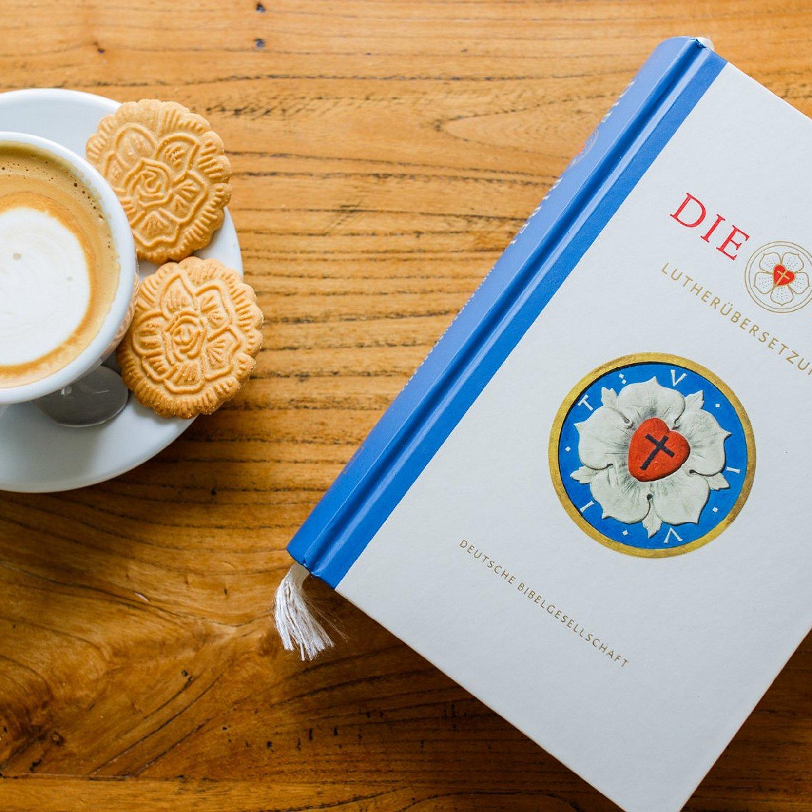 Bibel Lutherbibel neben einer Kaffeetasse mit Milchschaum in Apfelform und Keksen in Form der Lutherrose