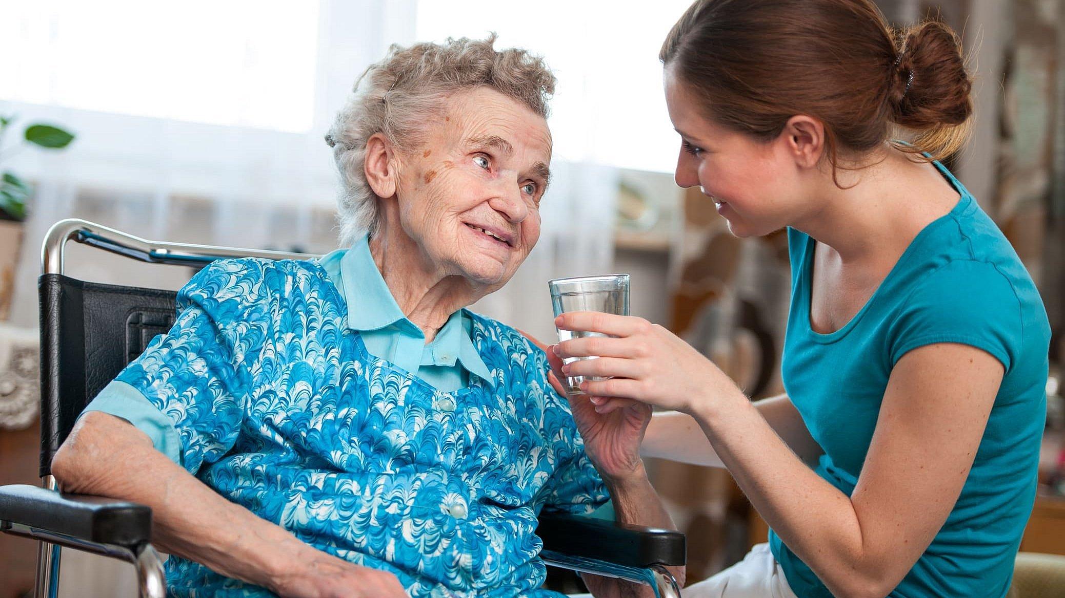 Eine alte Dame mit grauen Haaren sitzt im Rollstuhl eine junge Frau mit braunen Haaren sitzt ihr gegenüber und gibt ihr etwas zu trinken. Beide lächeln sich vertraut an.