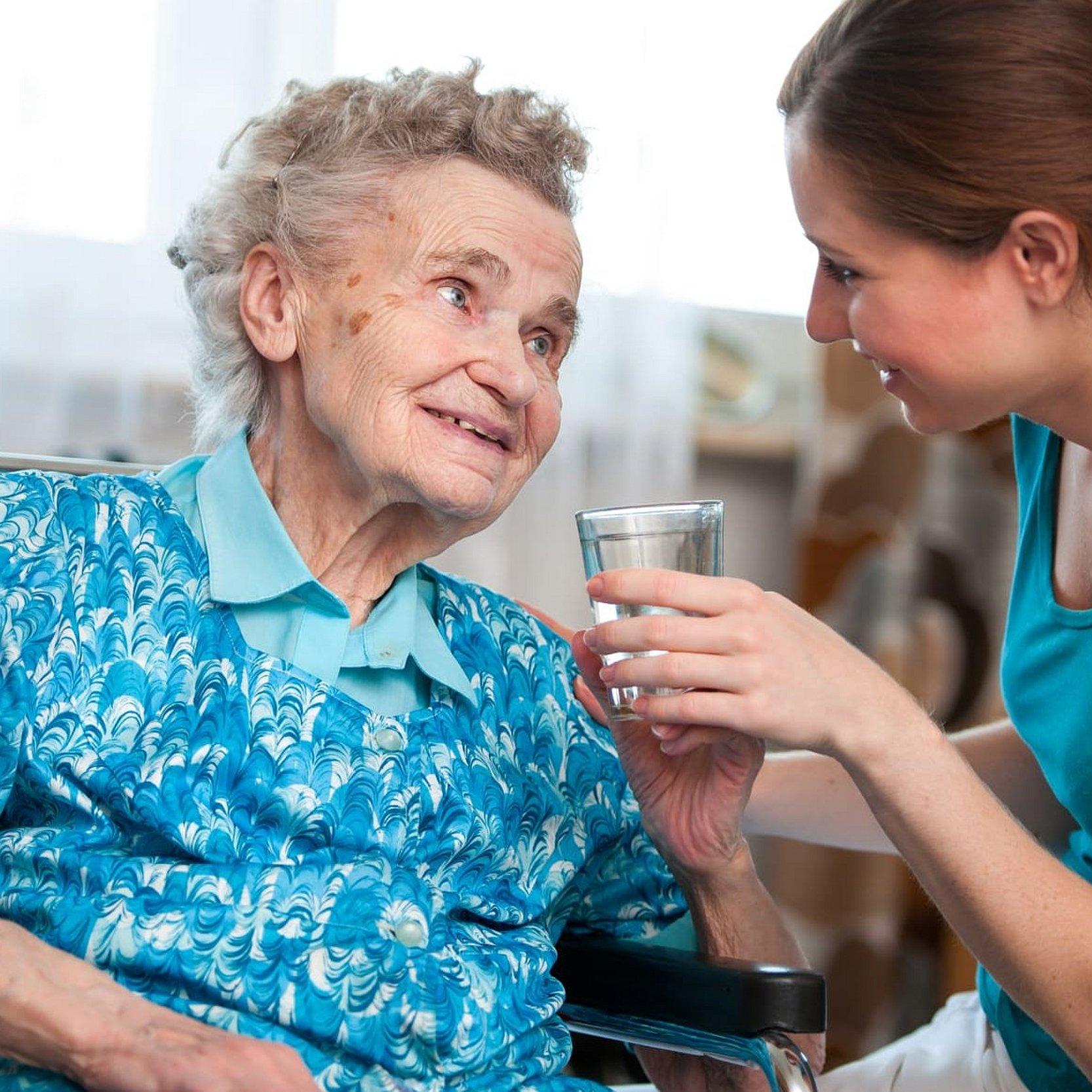Diakoniestation ambulante Pflege alter und kranker Menschen Eine alte Dame mit grauen Haaren sitzt im Rollstuhl eine junge Frau mit braunen Haaren sitzt ihr gegenüber und gibt ihr etwas zu trinken. Beide lächeln sich vertraut an.