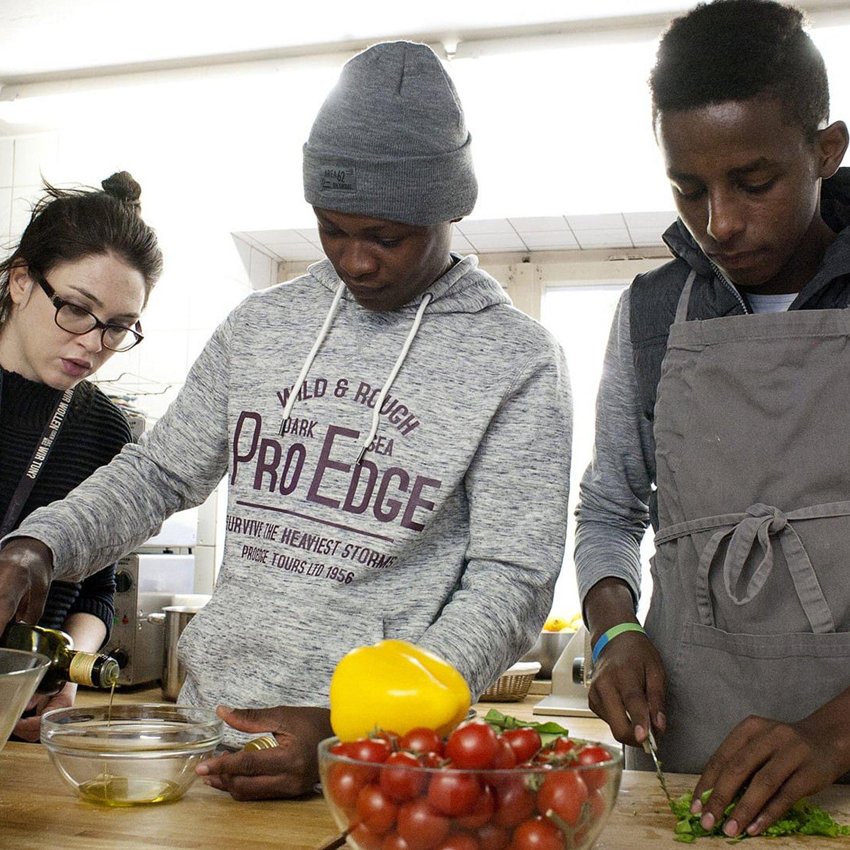 Flüchtlinge kochen mit junger Frau Zwei junge Geflüchtete kochen gemeinsam mit einer jungen Frau im Vordergrund steht eine Schüssel mit Tomaten.