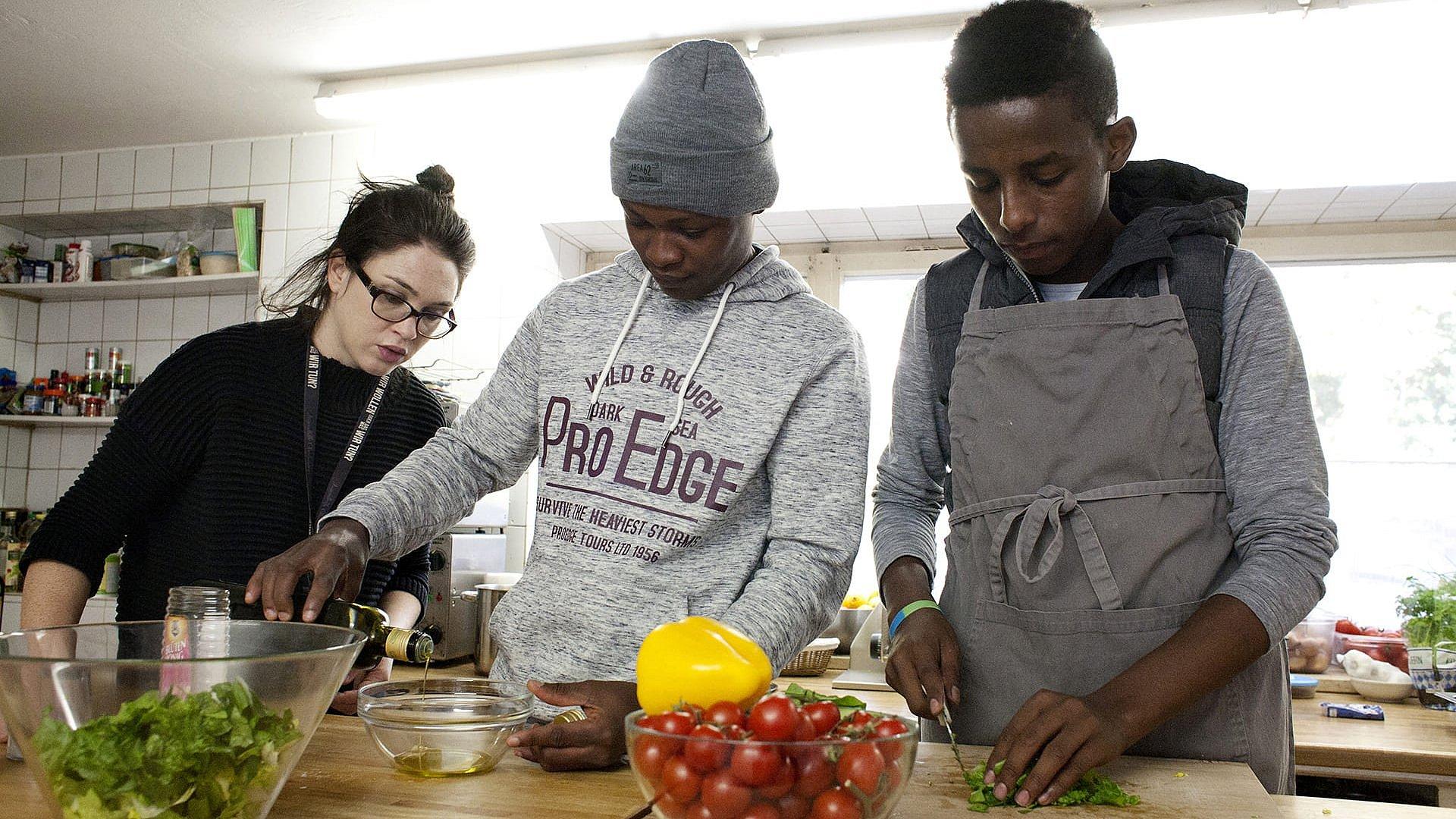 Zwei junge Geflüchtete kochen gemeinsam mit einer jungen Frau im Vordergrund steht eine Schüssel mit Tomaten.