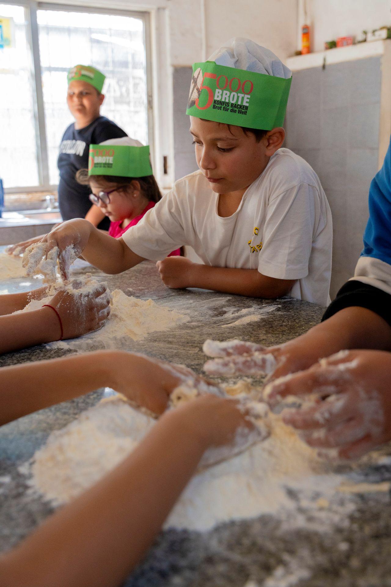 Brotback-Workshop Kinder kneten gemeinsam Teig auf einem mit Mehl bestäubten Tisch. Im Vordergrund ist ein Junge mit einer grünen Backmütze zu sehen, im Hintergrund weitere Kinder beim Backen.