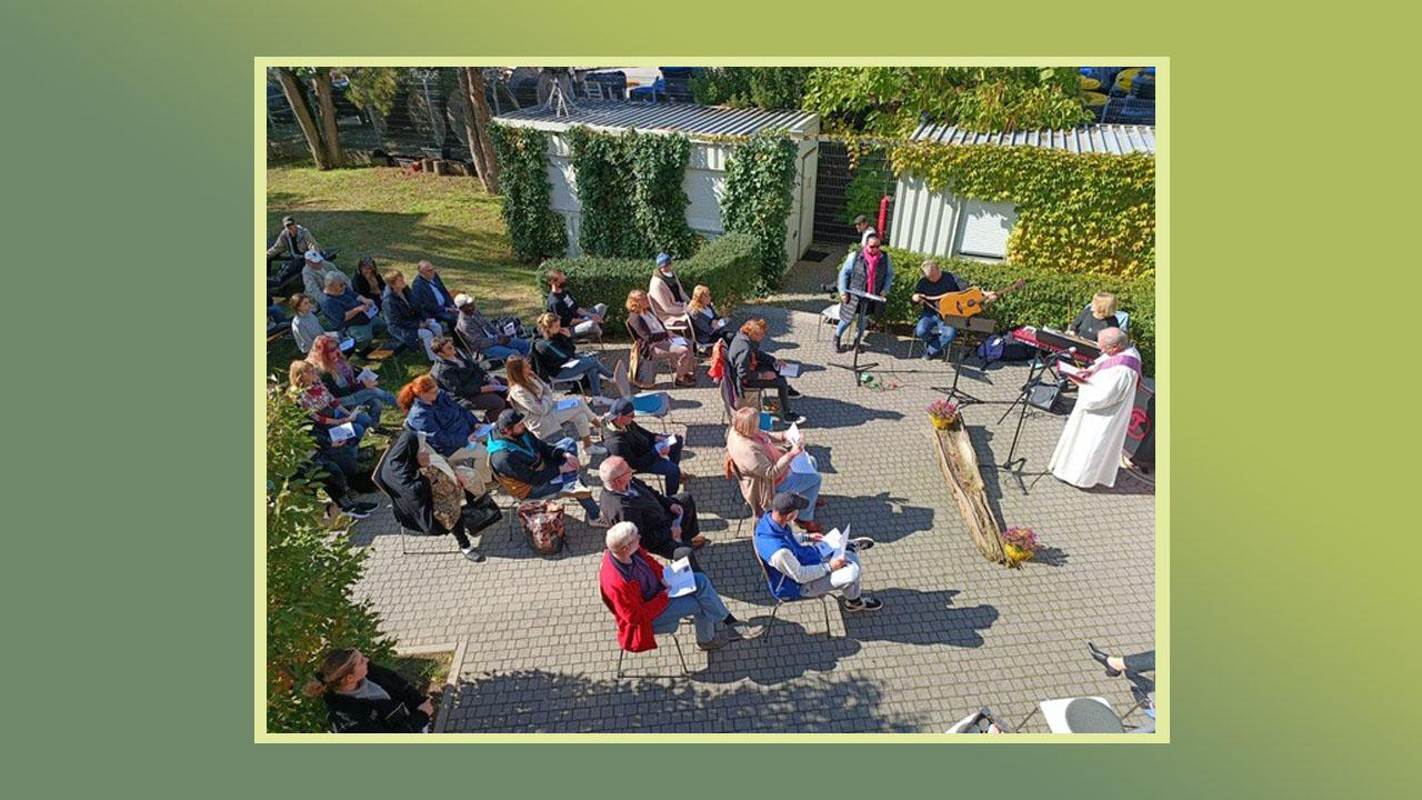 Gedenkgottesdienst  Menschen sitzen draußen im Grünen, vorne stehen ein Pfarrer im schwarzen und einer im weißen Talar