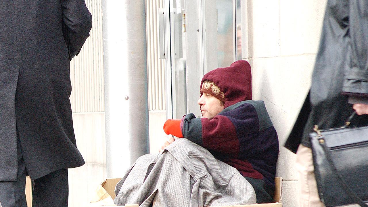 Obdachloser Mann sitzt im Winter in einer Fußgängerzone, Menschen eilen vorbei