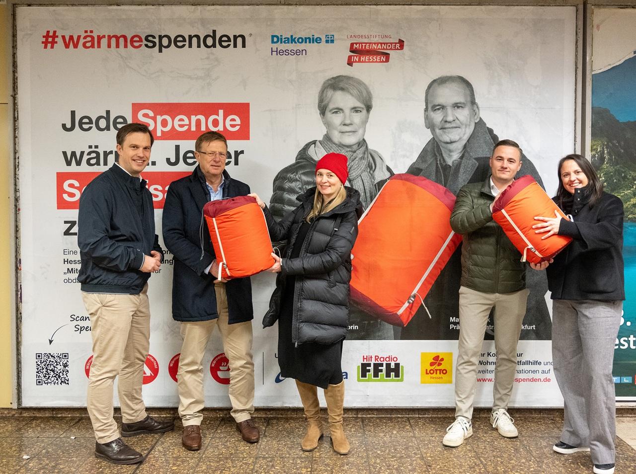 Aktion #Wärmespenden Gruppenfoto vor Straßenplakat. Unterstützer*innen mit den roten Schlafsäcken