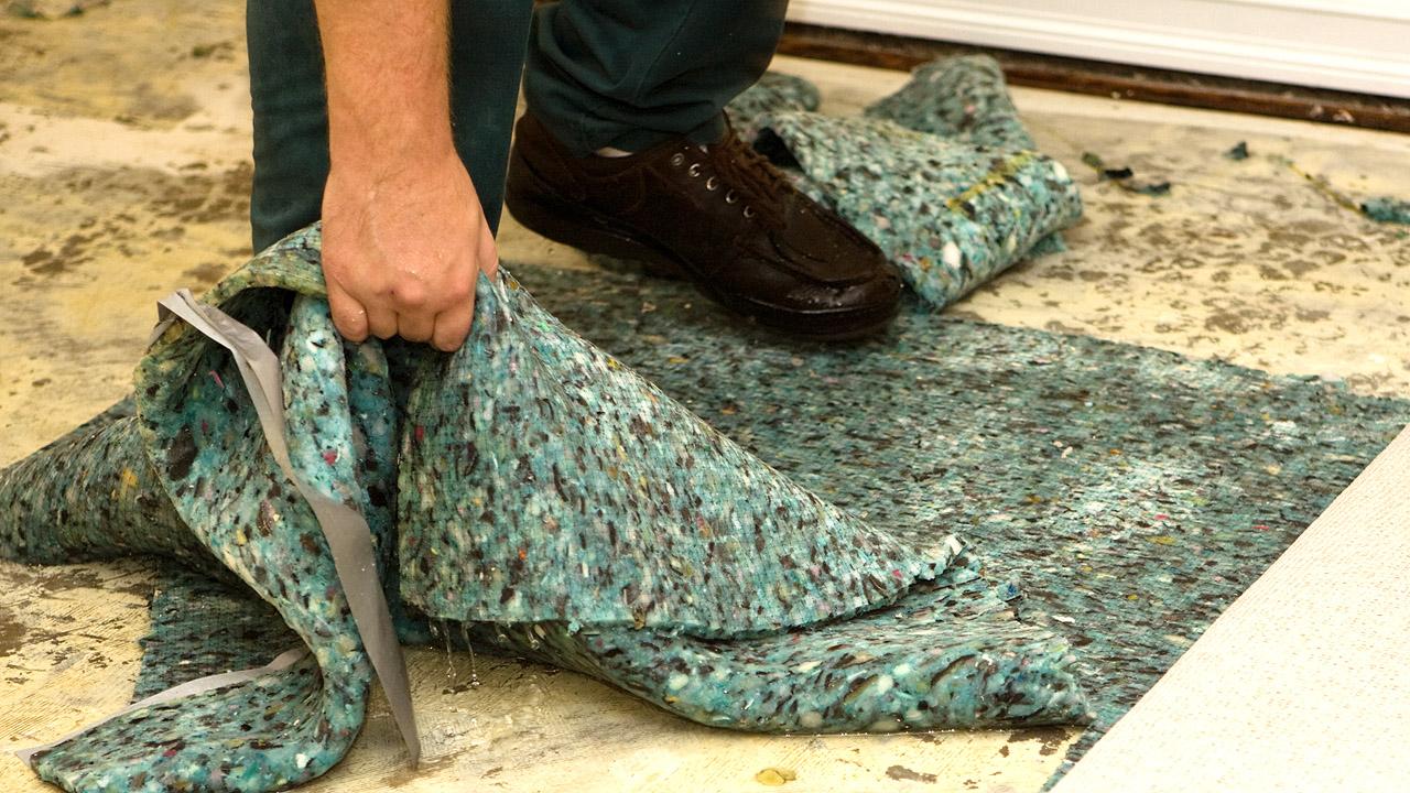 Eine Hand zieht einen nassen Teppichboden vom schmutzigen Untergrund