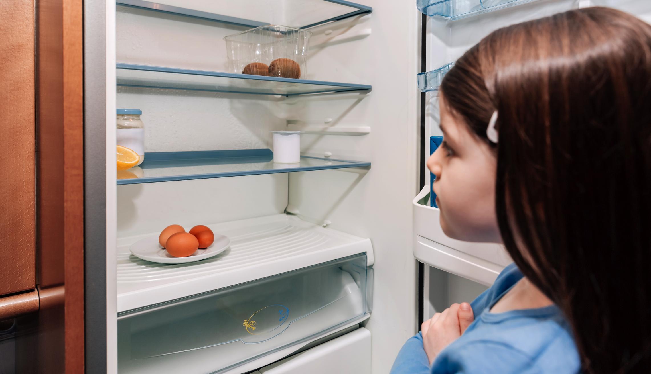 Mädchen schaut in fast leeren Kühlschrank.