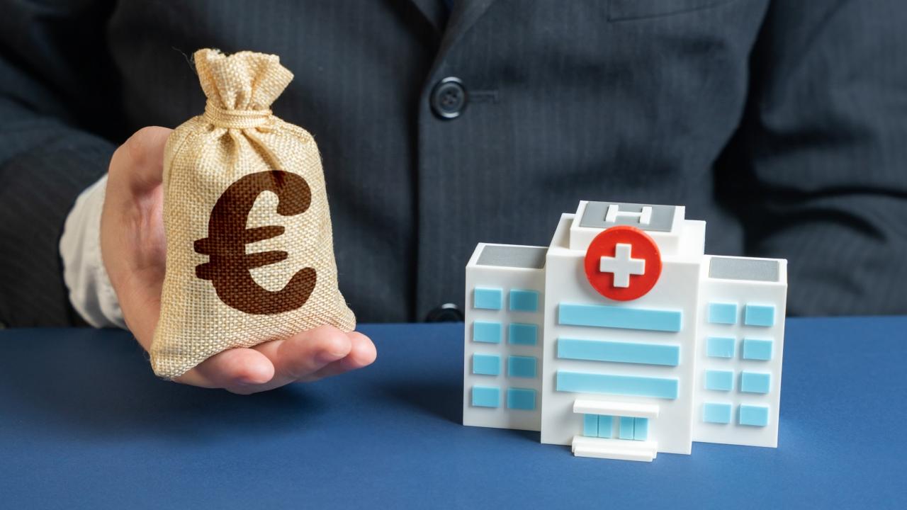 Modell eines Krankenhauses und Mann mit kleinem Geldsack, auf dem ein Euro-Zeichen ist