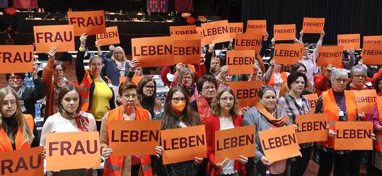 Frauen demonstrieren mit orangefarbenen Schildern auf der EKHN Synode