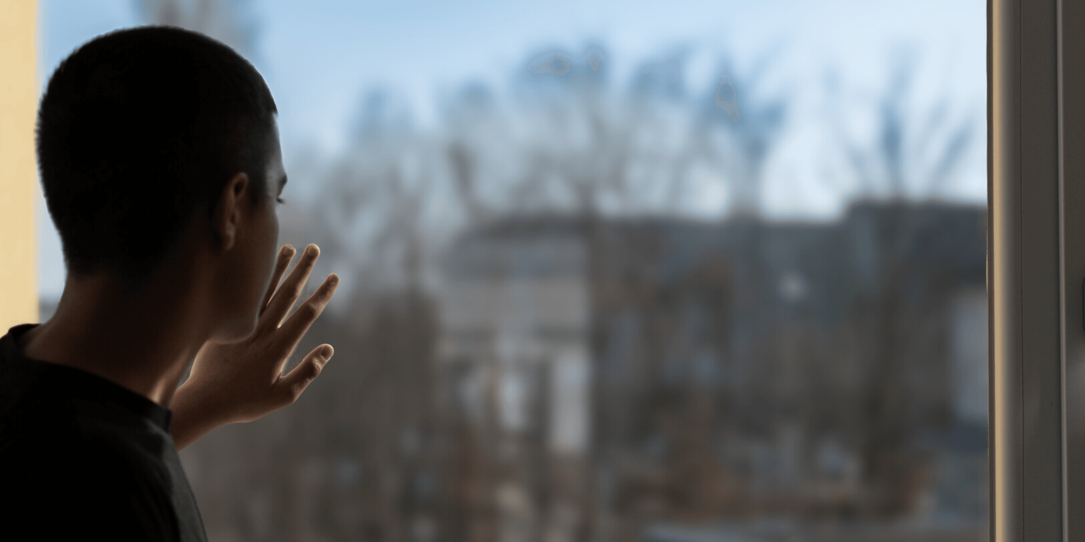 Junger Mann schaut aus dem Fenster Ein Mann steht am linken Rand alleine am Fenster einer Flüchtlingsunterkunft und schaut auf ein entferntes Gebäude, dass durch Bäume nicht ganz sichtbar ist