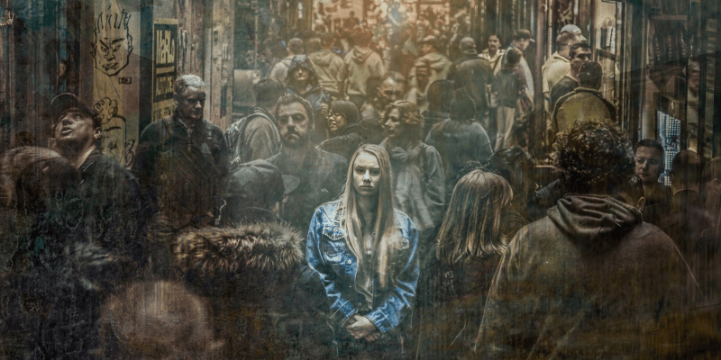 Traurige Frau in einer Menschenmenge eine blonge mit Jeansjacke bekleidete Frau befindet sich zentral in einer Menschengruppe in einer vollen Straße umgeben von Häuser und Geschäften. Das Bild wirkt wie eine Zeichnung. Die Frau ist hell und erstrahlt. Der Rest sehr dunkel.