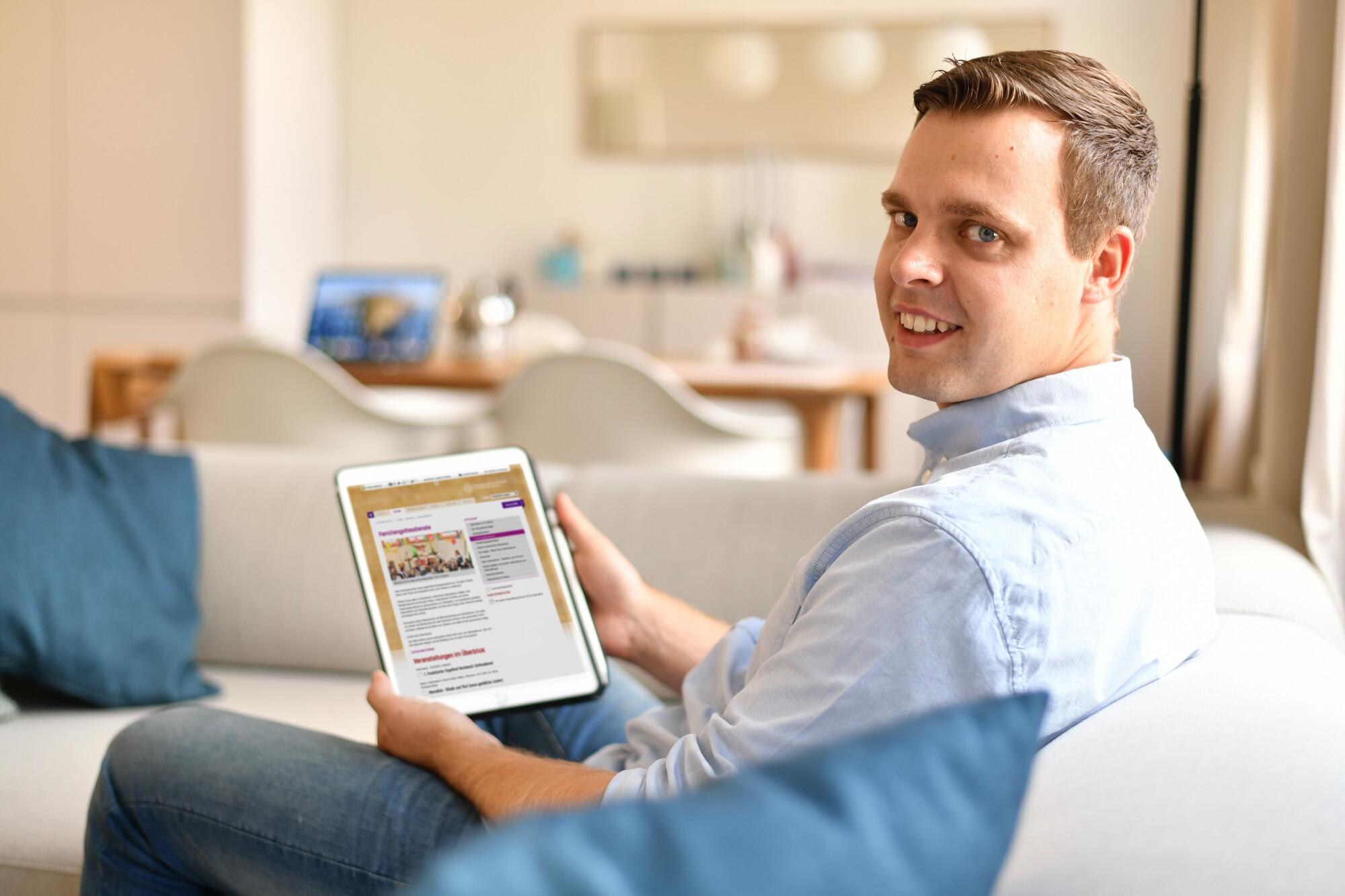 Ein Mann - aufgenommen von seitlich hinten - sitzt auf einer Couch und schaut in Richtung Kamera. Im unscharfen Bildvordergrund sind Sofakissen und im unscharfen Hintergrund ist ein Küchentisch mit Stühlen zu sehen.