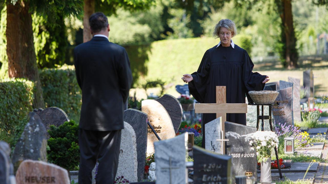 Eine evangelische Pfarrerin am Grab zwischen vielen Grabsteinen. Ein Mann steht im Vordergrund
