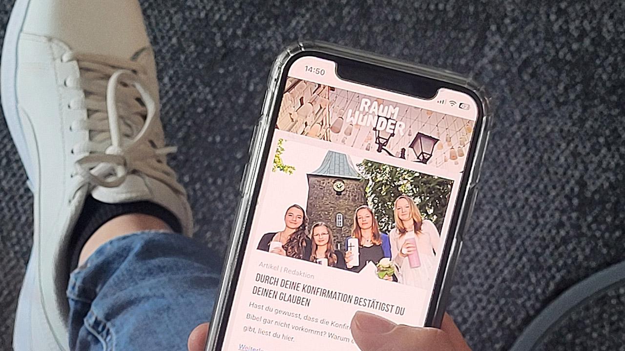 Konfiweb Hände einer Jugendlichen halten ein Smartphone, auf dem die Konfiweb-Startseite zu sehen ist