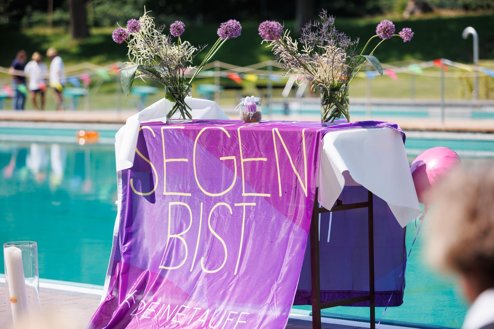 Tauffest im Naturschwimmbad Höhr-Grenzhausen: Eine Fahne der Taufinitiative der EKD liegt auf einem Tisch und hängt bis zum Boden. Der Tisch steht vor einem Schwimmbecken und ist mit Blumen und Taufkerzen dekoriert. Aufnahme seitlich von rechts.