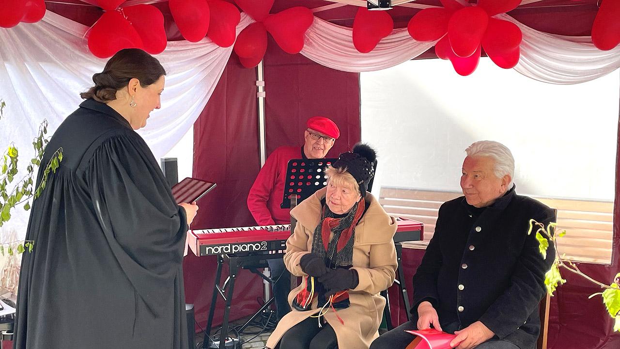 Trauung-to-go - Traupavillon Eine Pfarrerin im Talar steht vor einem älteren Paar, das sitzt. Unter der Decke hängen rote Herz-Luftballons.