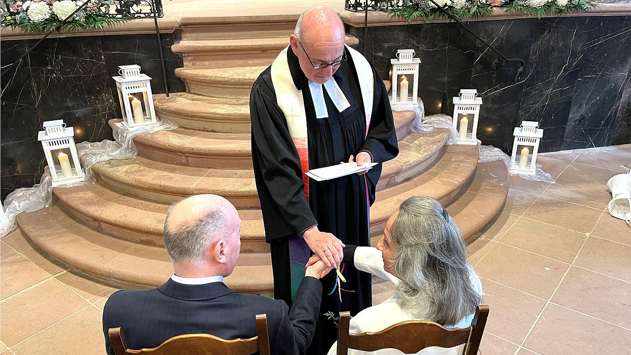 Trauung-to-go Ein Pfarrer steht vor einem älteren Paar. Dahinter ist eine Treppe, auf der an den Seiten weiße Laternen stehen.