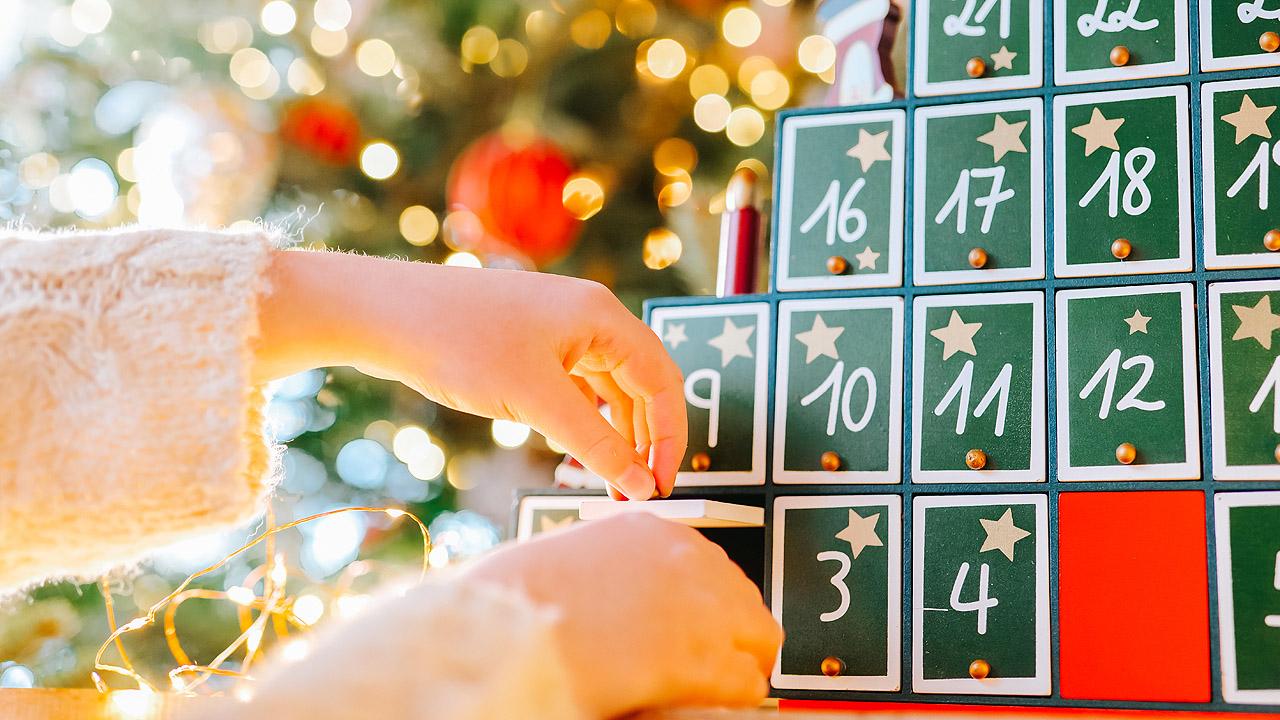 Die Hand eines Mädchen öffnet eine kleine Schublade eines bemalten Adventskalenders aus Holz