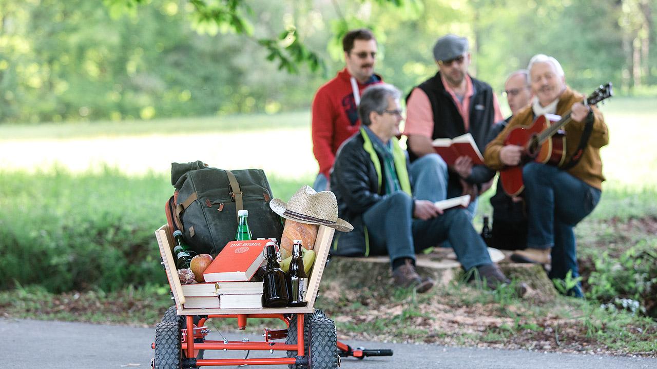 Im Vordergrund ein Bollerwagen mit Bibel, dahinter musizierende Männer mit Gitarre