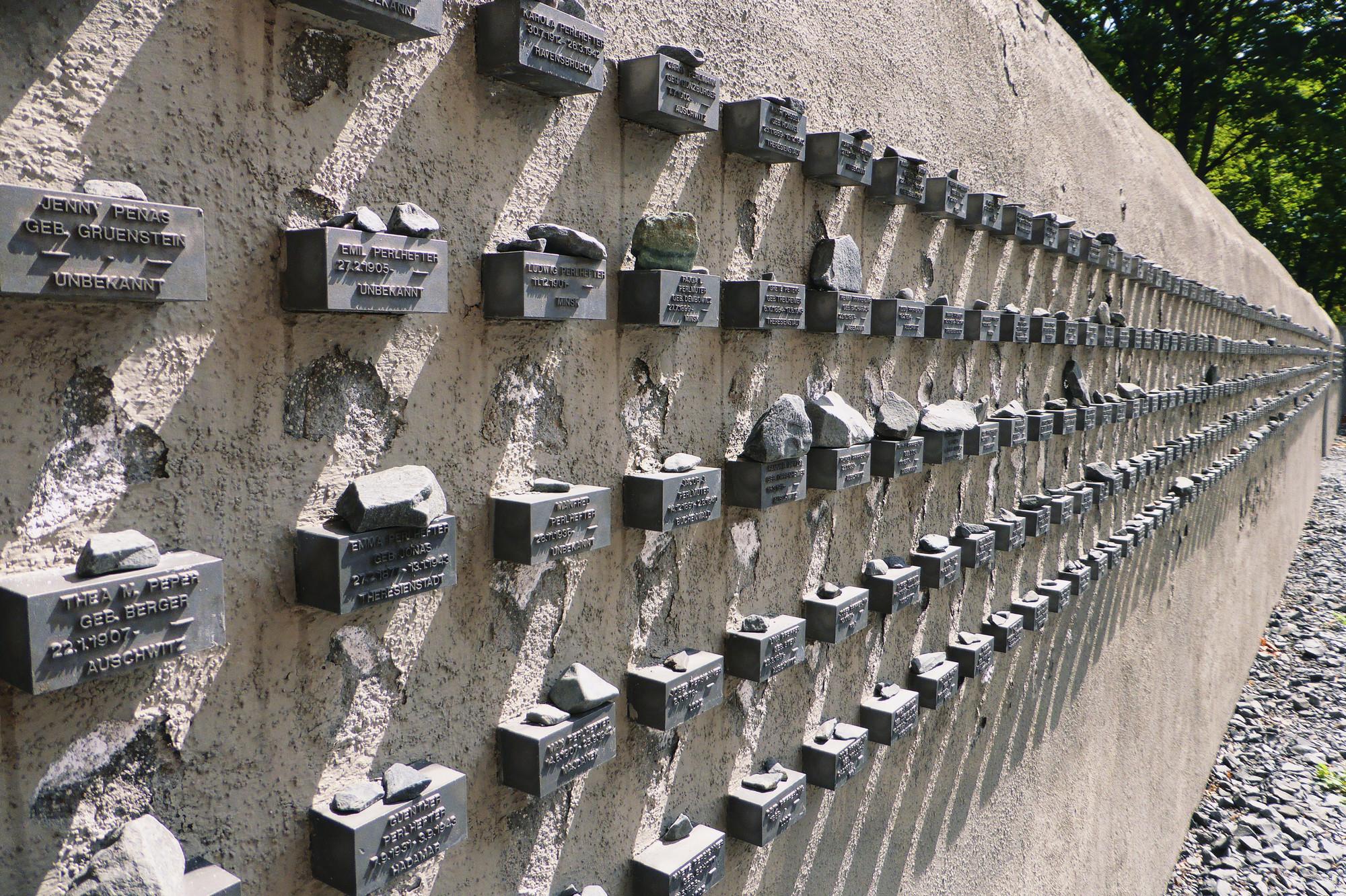 Zu sehen sind Gedenktafeln an der Mauer des jüdischen Friedhofs Battonstraße in Frankfurt/M für die aus der Stadt deportieren jüdischen Menschen. Seitlich an der Mauer befinden sich kleine Boxen, in welche Namen sowie Geburts- und Todesdaten eingraviert sind. Auf diesen Boxen liegen kleine Gedenksteine.