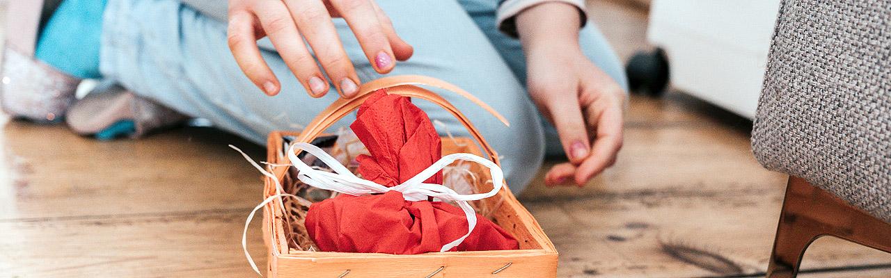 Ostereiersuche Kind rutscht auf Knien hinter ein Sofa, in einem Körbchen liegt ein kleines, verpacktes Geschenk