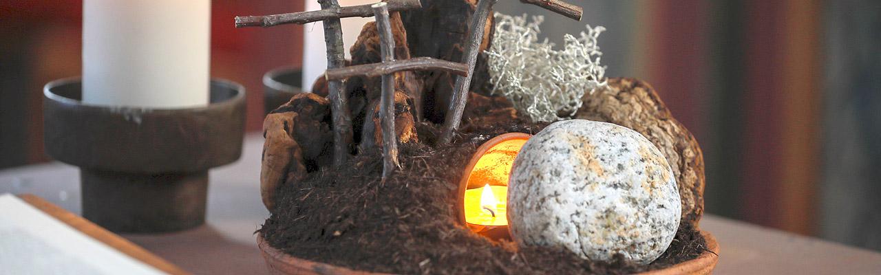 Osterkrippe Körbchen mit eine Höhle, in der eine Kerze brennt, davor der weggerollte Stein, dahinter Kreuze aus Zweigen