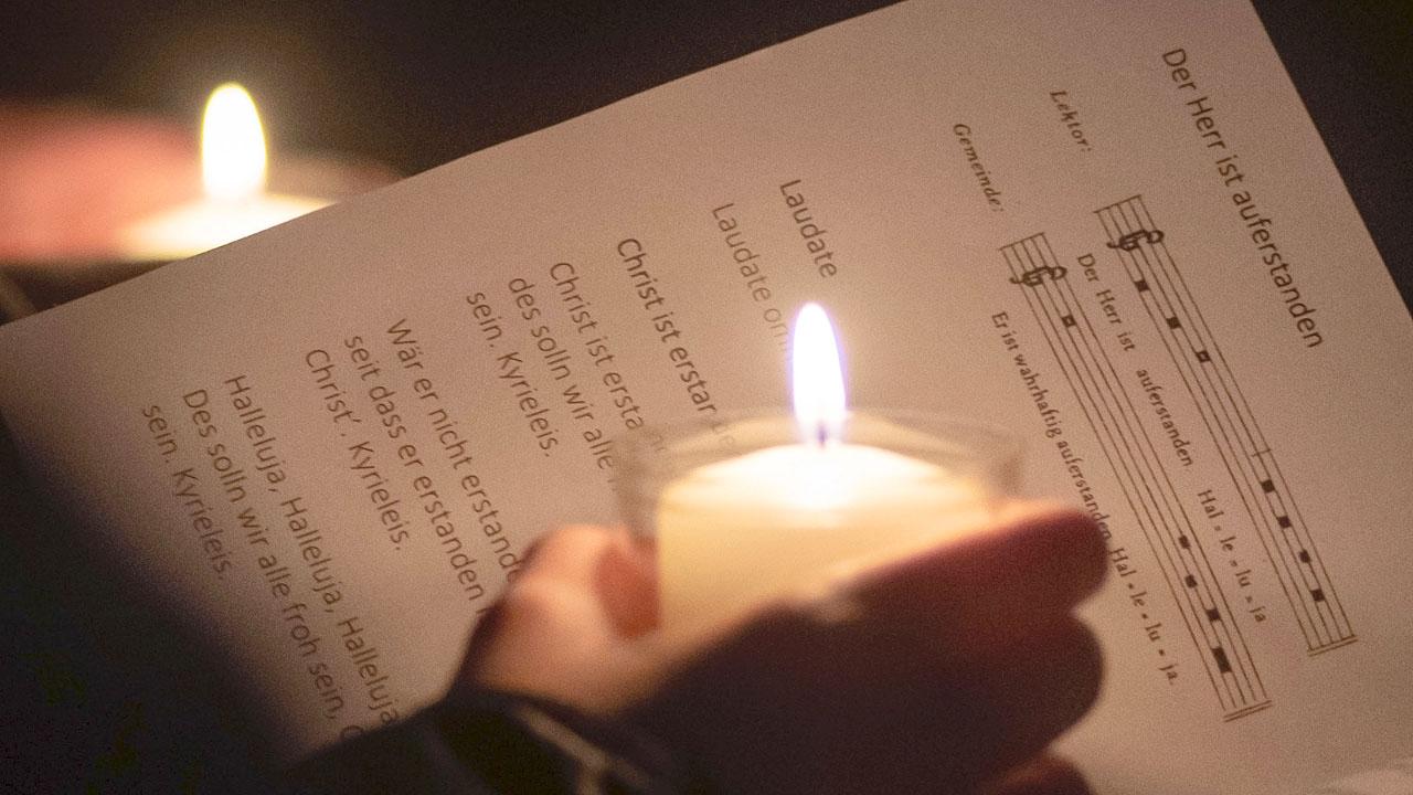Osterlicht im Gottesdienst Eine Hand hält eine Osterkerze, dahinter sind Noten von Lieder zu sehen, die erhellt werden