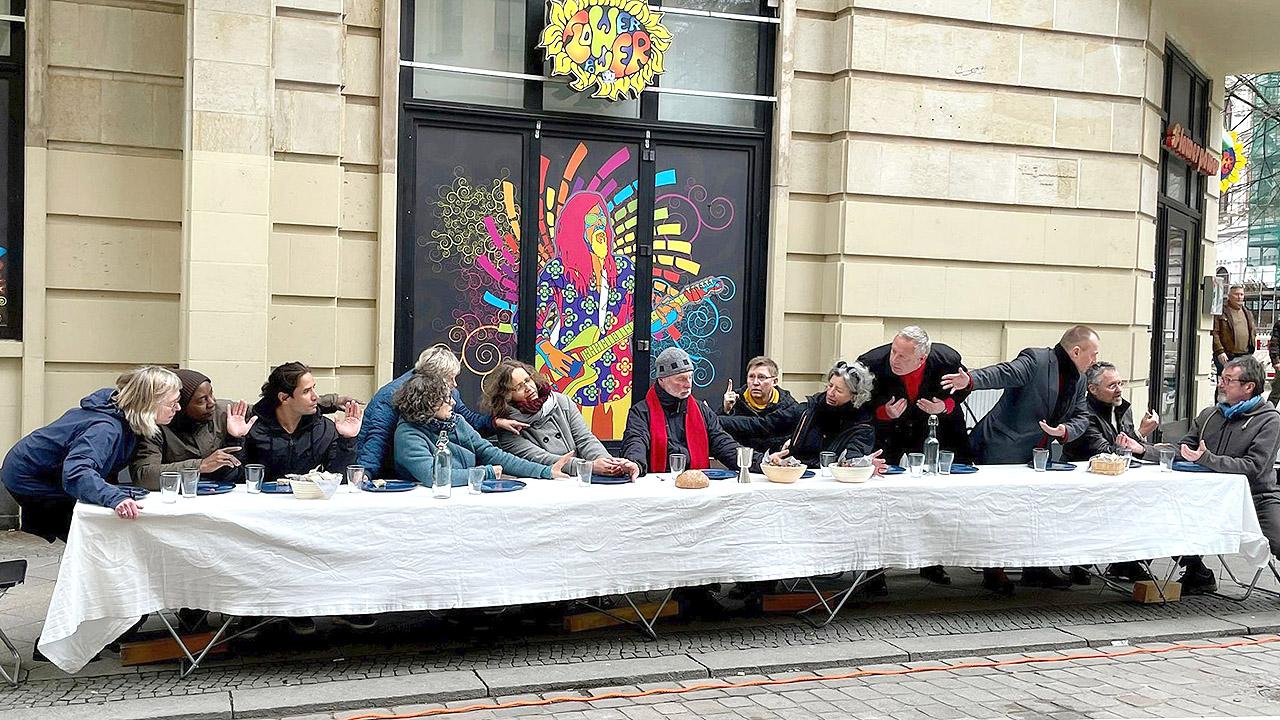 Menschen in Alltagskleidung an einem langen Tisch auf einem Bürgersteig