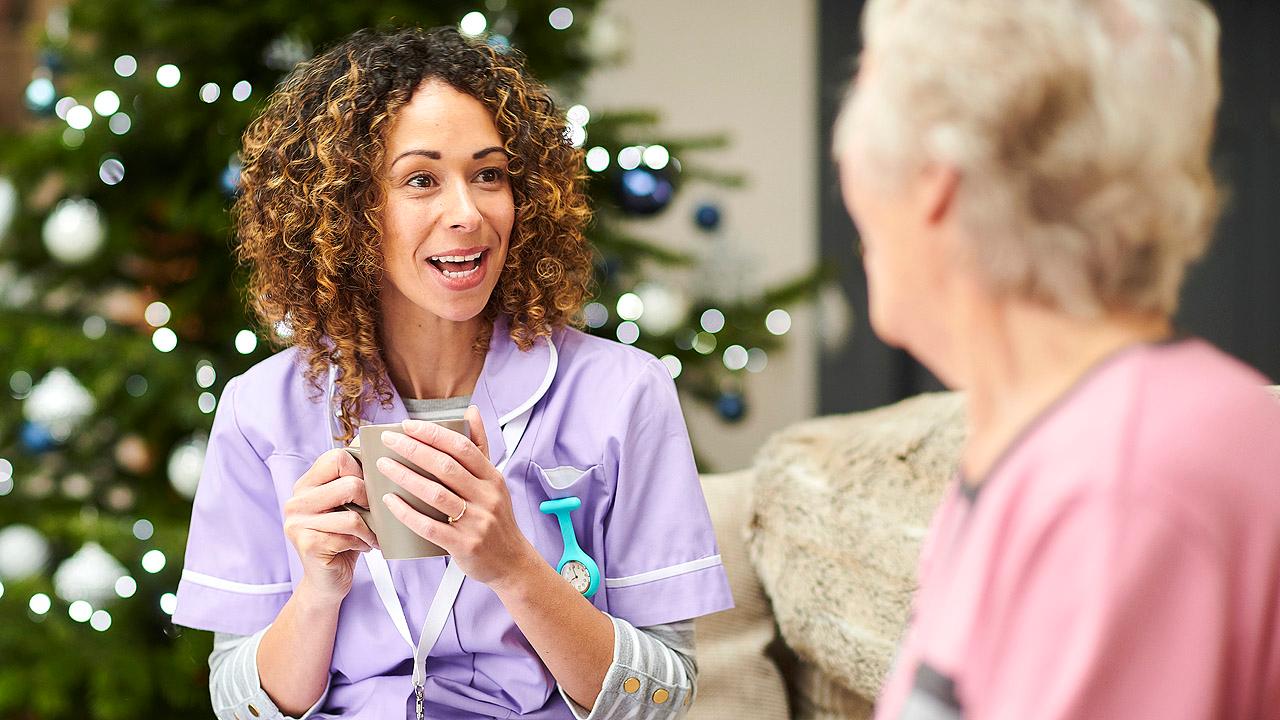 Pflegerin unterhält sich mit Seniorin, dahinter steht ein Weihnachtsbaum