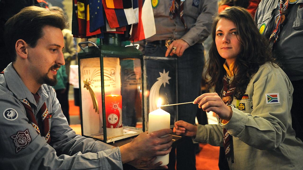 Eine junge Frau in Pfadfinder-Uniform zündet eine Kerze an, dahinter steht eine Kerze in einem gläsernen Windlicht