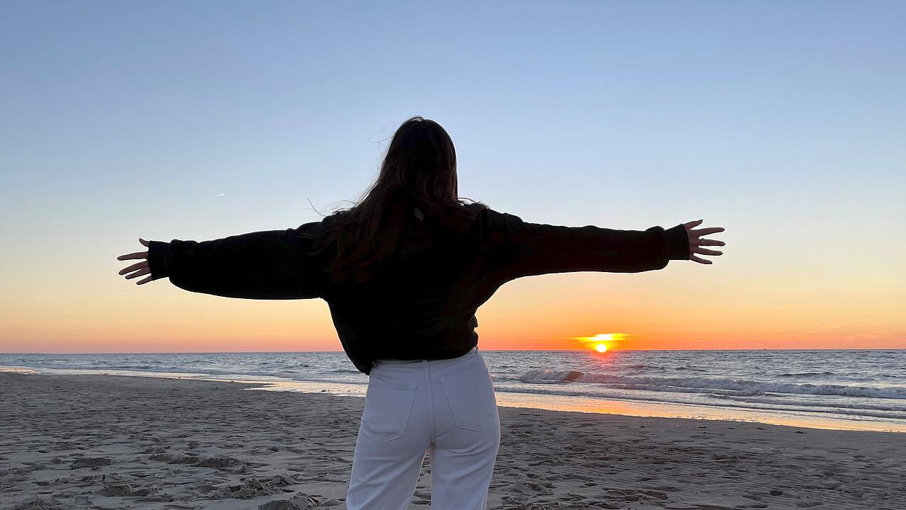 Junge Frau breitete ihre Arme auf und schaut in den Sonnenuntergang am Meer