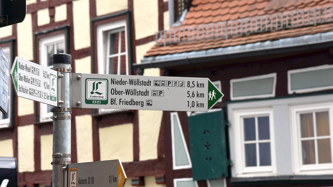 Lutherweg in der Wetterau Schild mit Lutherweg-Logo, im Hintergrund Fachwerkhäuser