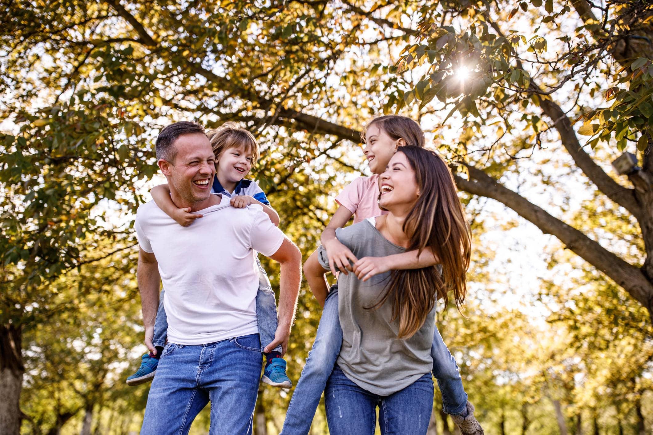Familie hat Spaß zusammen und wandert Ein Vater und eine Mutter gehen durch den Wald, sie lachen und haben jeweils ein Kind auf ihrem Rücken. Die Kinder lachen und schauen sich an. Alle sind glücklich beim Huckepack-Spiel.