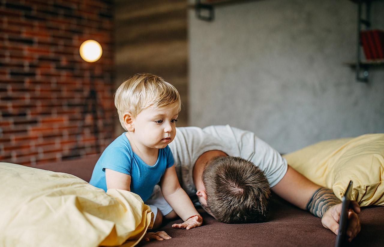 Kopf eines Vaters liegt auf einem Bett, daneben ein Kleinkind