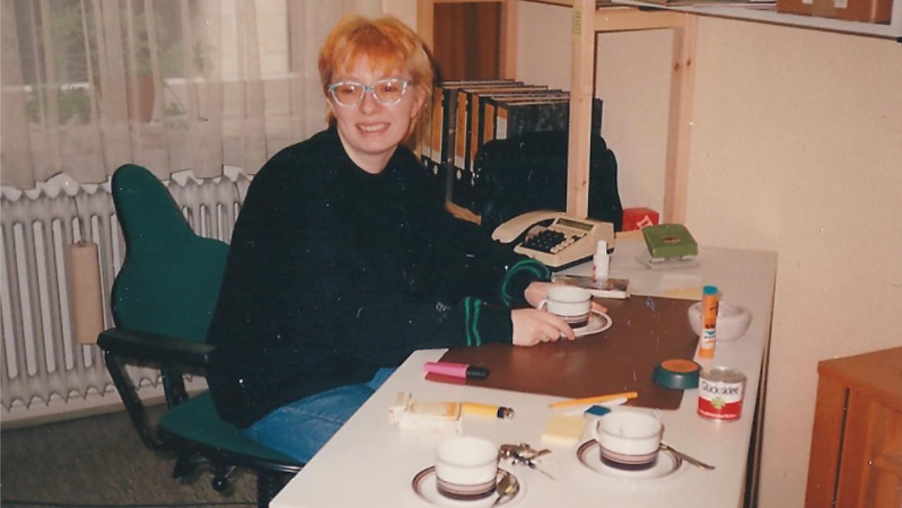 Antje Schrupp 1989 Antje Schrupp sitzt an einem Schreibtisch auf dem drei Tassen stehen, sie sitzt auf einem blauen Bürostuhl, im Hintergrund steht ein Bücherregal