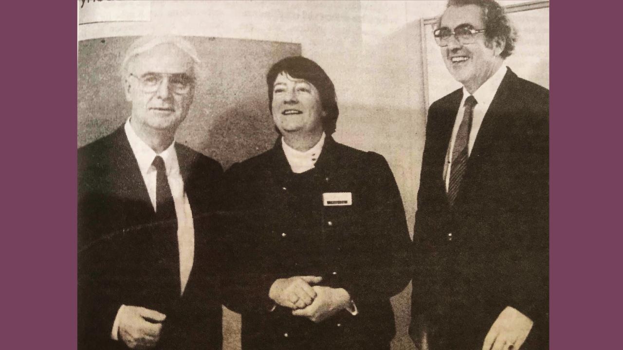 Helga Trösken mit Propst Trautwein und Kirchenpräsident Spengler Helga Trösken im schwarzen Kostüm mit weißem Kragen steht zwischen zwei Männern, die beide einen dunklen Anzug und Krawatten tragen.