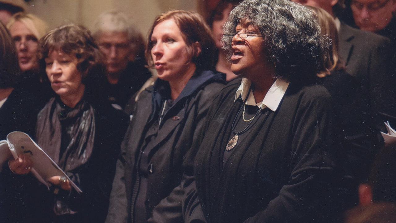 Flois Knolle Hicks In dunkler Kleidung singen Frauen und Männer andächtig in einem Raum