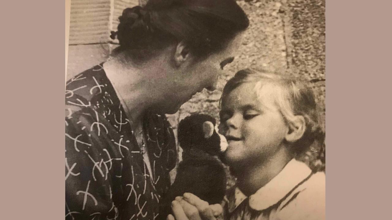Katharina Staritz und Kind Katharina Staritz mit einem gemusterten Kleid, die dunklen Haar zu einem Dutt gesteckt hat ein Stofftier in der Hand neben ihr steht ein Mädchen mit blonden Haaren und weißer Bluse, die zum Stofftier blickt