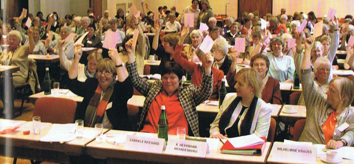 Ein großer Saal mit vielen Frauen, die an Tischen sitzen, wie auf einer Konferenz und mit erhobenden Händen abstimmen