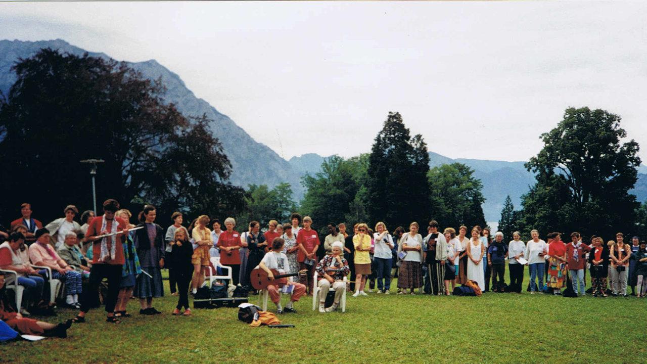Frauensynode 1992 in Österreich Eine große Gruppe von Frauen steht im Halbkreis auf einer Wiese, in der Mitte spielt eine Frau Gitarre im Sitzen. Im Hintergrund Berge