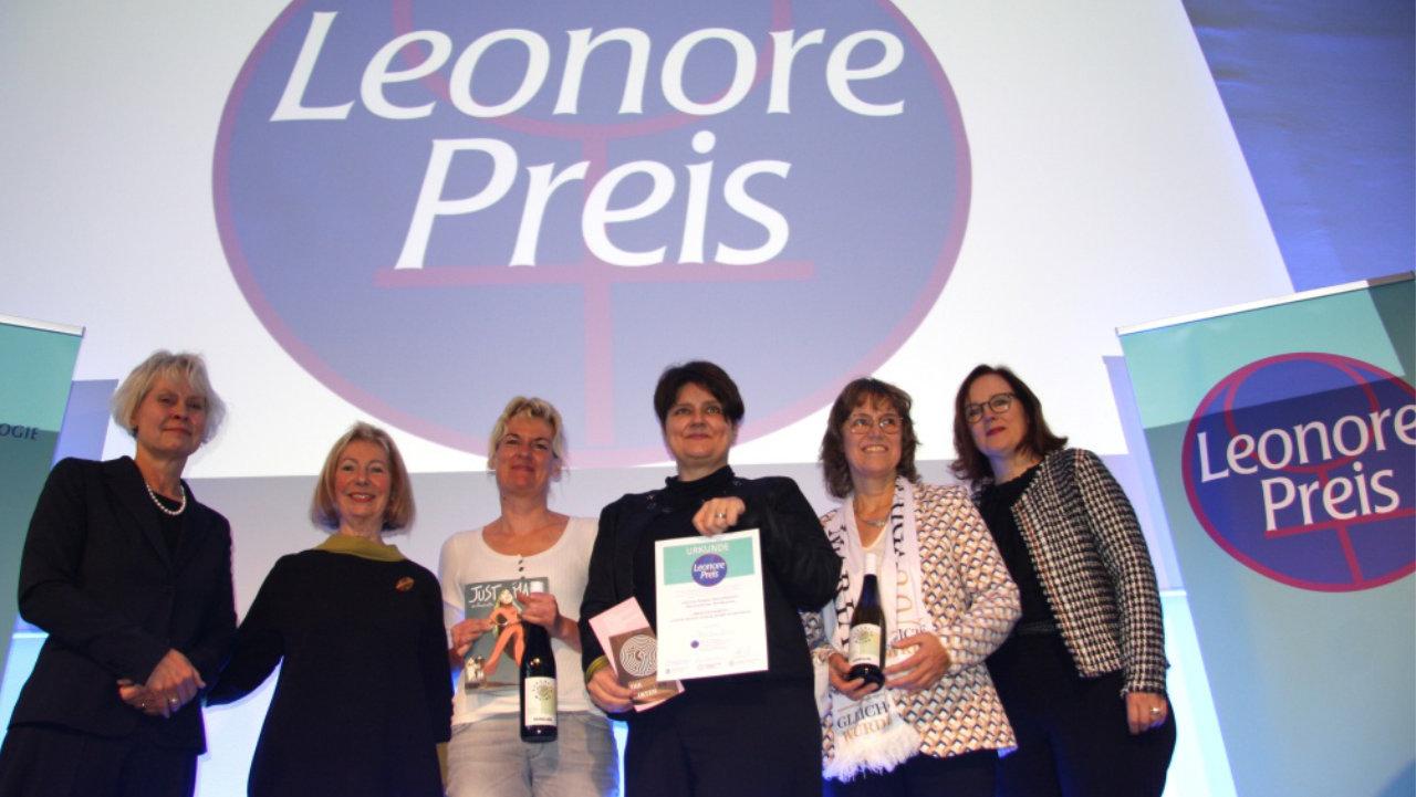 Preisverleihung 2023 Sechs Frauen auf einer Bühne, die mittlere Person hält die Urkunde in der Hand im Hintergrund ist das Logo des Leonore-Preises zu sehen.